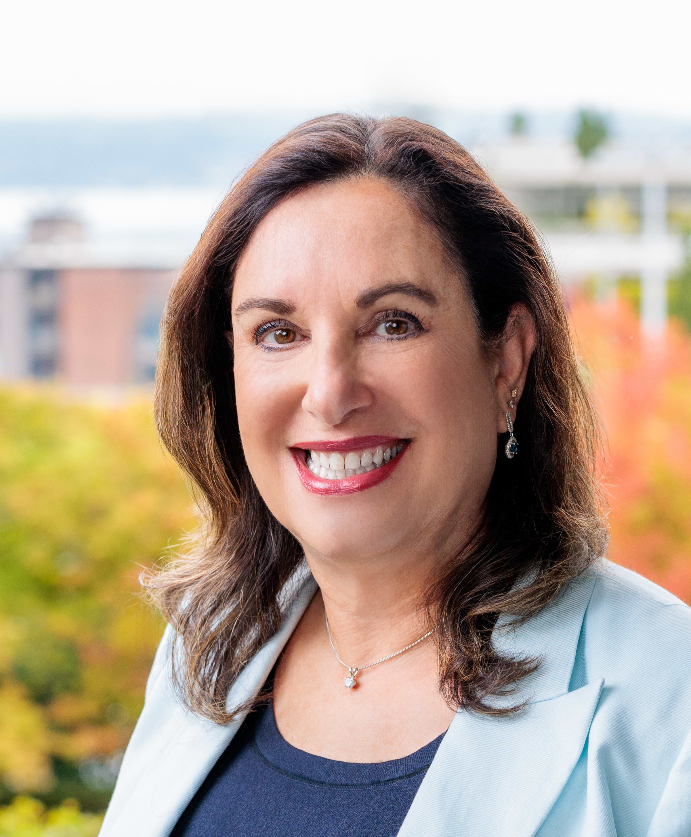 Sigal Zarmi with dark hair stands outdoors, wearing a light blue blazer. Background shows blurred trees and buildings.